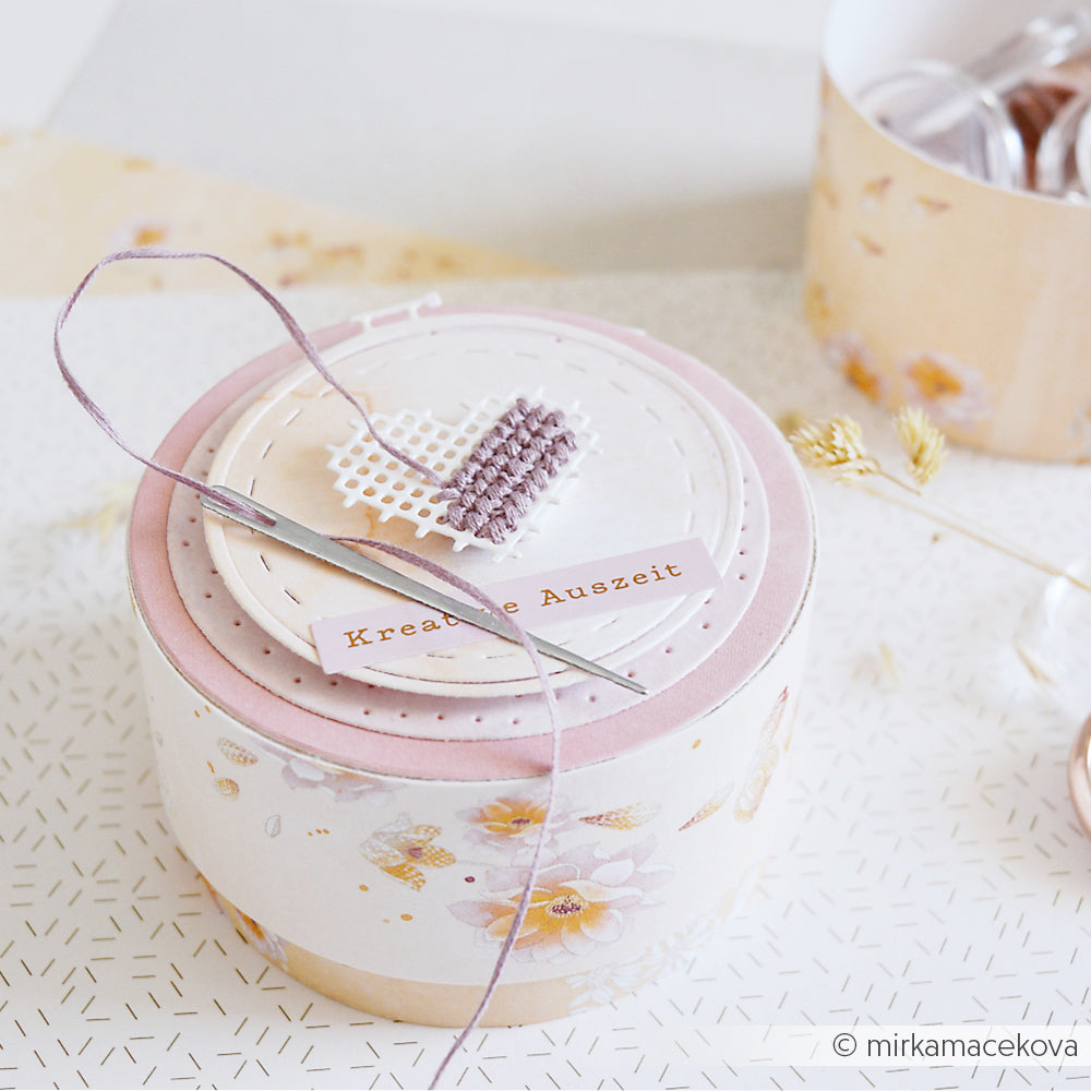 Runde Geschenkbox von Alexandra Renke mit Blumenmuster, Nadel, Faden und einem Kreuzstich-Herz auf dem Deckel – ideal als Geschenk oder in Kombination mit den „Motivstanze Stickrahmen Rund“-Stanzformen für eine persönliche Note.