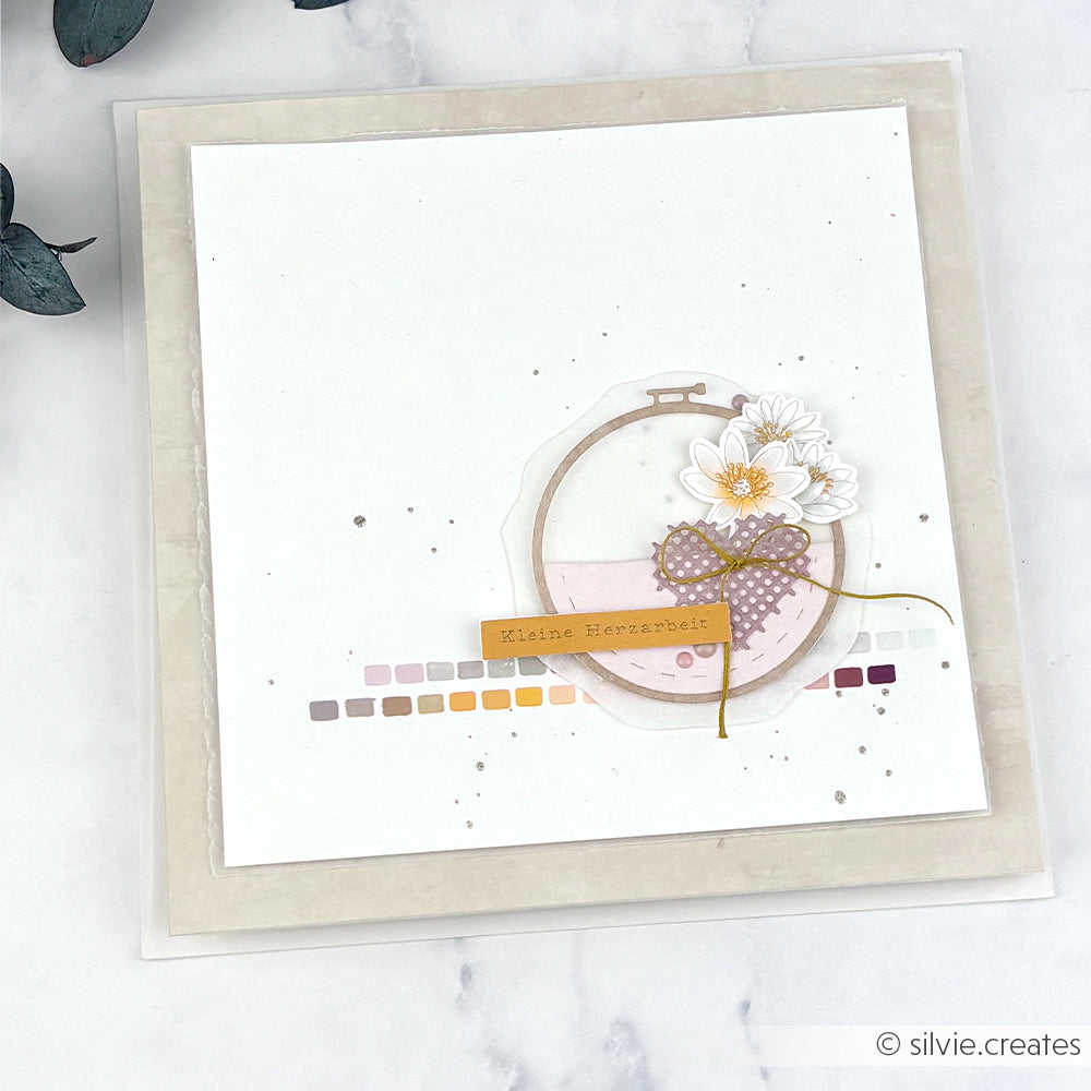 Handgefertigte Grußkarte von Alexandra Renke mit Blumen- und Schleifenmotiv, dem deutschen Text „Kleine Herzarbeit“ sowie Akzenten aus Designpapieren und der Motivstanze „Stickrahmen Rund“ von DieCut Systeme auf schlichtem weißem Hintergrund.