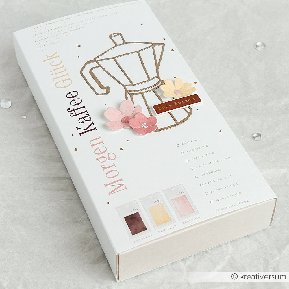 Geschenkbox „Weißer Kaffee“ von Alexandra Renke mit einer Illustration der Espressokanne von Motivstanze, rosa Blumen und dem deutschen Text „Morgen Kaffee Glück“.