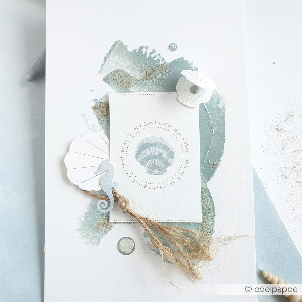 Handgefertigte Karte von Alexandra Renke mit einem Muschelmotiv aus blauen und beigen Designpapieren, Muscheln, Schnur und Akzenten aus dem Motivstanz-Set „Falt-Muschel“ auf weißem Hintergrund.