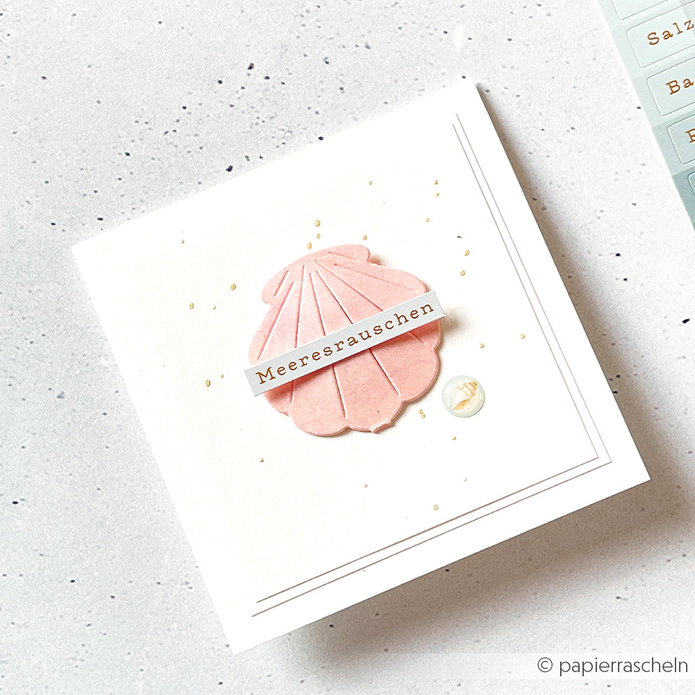 Quadratische Karte mit einer rosa Muschel, einer kleinen Muschel und dem deutschen Wort „Meeresrauschen“ auf weißem Hintergrund, gestaltet mit dem „Motivstanze Falt-Muschel“-Set von Alexandra Renke – elegante Stanzformen für Designpapier.