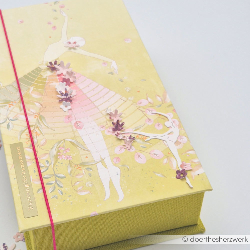 Ein gelbes Buch von Alexandra Renke mit einer Ballerina-Illustration, rosa 3D-Blumen aus Designpapier, die mit einem roten Gummiband befestigt sind.