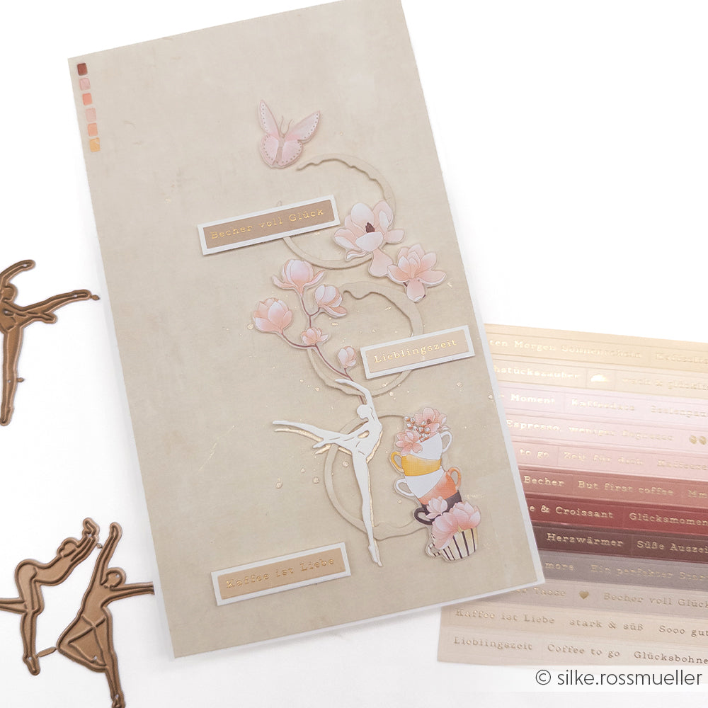 Eine handgefertigte Karte von Alexandra Renke mit pastellfarbenen Blumen, einem Schmetterling und übereinander gestapelten Teetassen auf beigem Premium-Papier, verziert mit goldenen Schriftzügen.