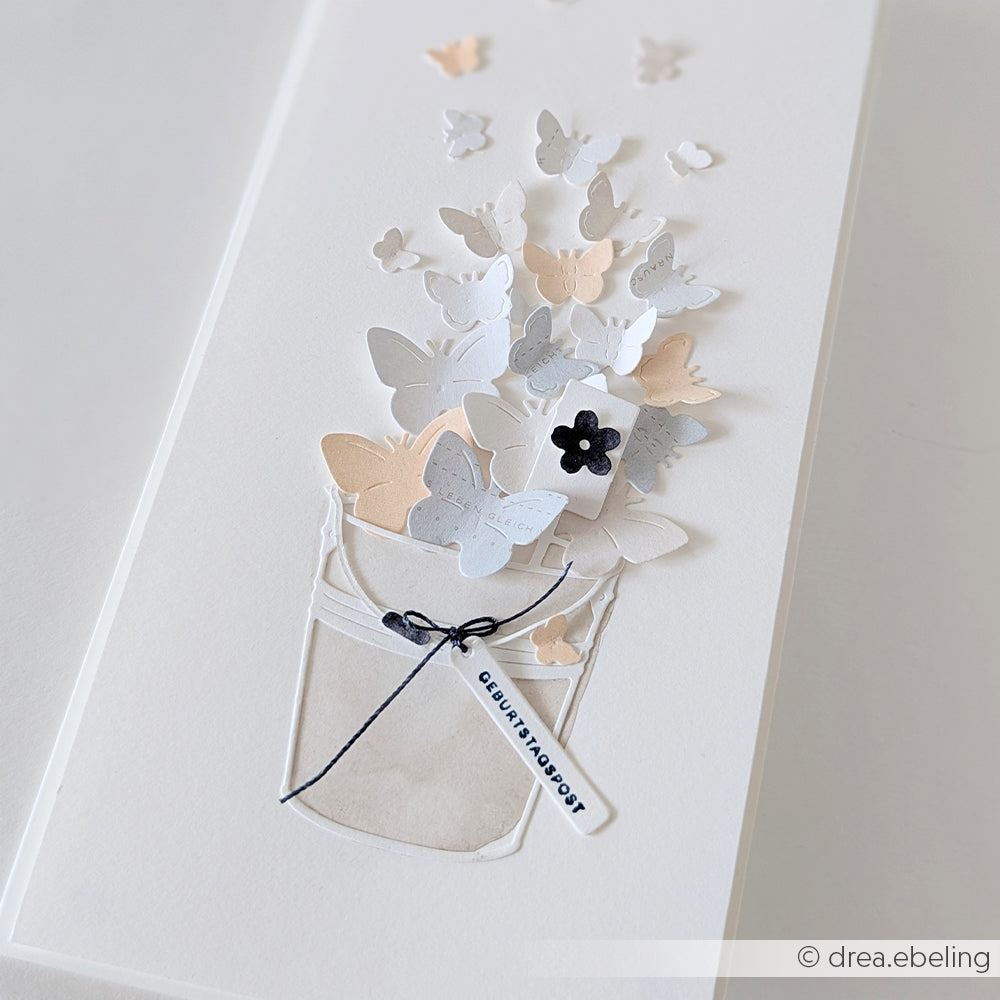 Handgefertigte Karte mit 3D-Papier-Schmetterlingen, die aus einem Eimer fliegen, gestaltet mit dem „Motivstanze Garteneimer“-Stanzsystem von Alexandra Renke und verziert mit einer schwarzen Schleife und einer kleinen Blumenverzierung – perfekt, um Ihre Lieblings-Stanzen für Designpapier in Szene zu setzen.