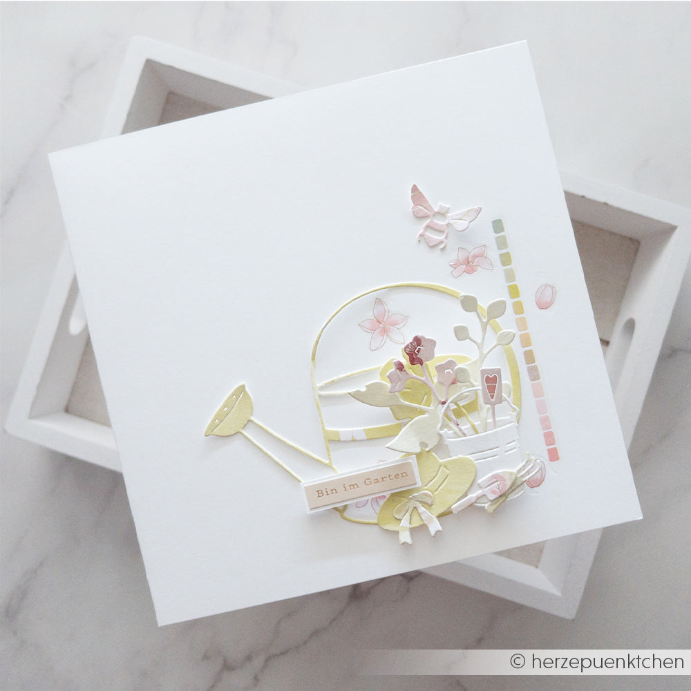 Quadratische weiße Karte mit pastellfarbenen Ausschnitten zum Thema Garten, darunter eine Motivstanze „Gießkanne“ und Blumen, die mit Designpapieren und Stanzsystemen von Alexandra Renke gestaltet wurden und der Karte eine zarte Note verleihen.