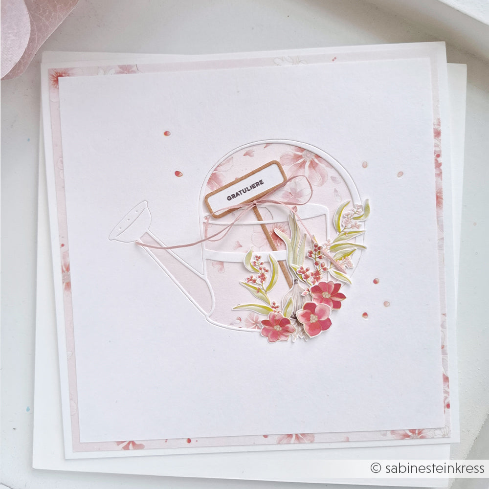 Handgefertigte Grußkarte von Alexandra Renke, hergestellt aus Designpapier und mit einer Stanze in Form einer Gießkanne, mit rosa Blumen und einem kleinen „Gratuliere“-Schild auf blumigem Hintergrund.