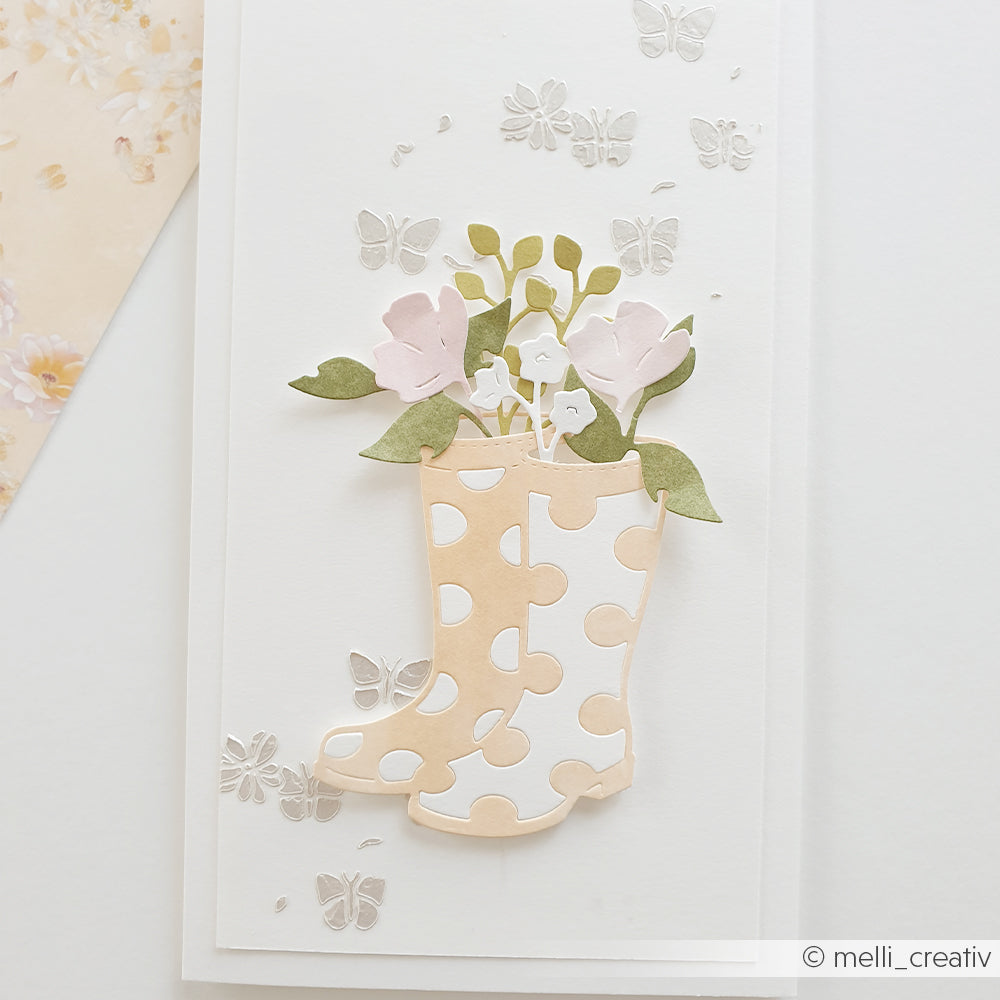 Die mit der „Alexandra Renke Motivstanze Gummistiefel“ gefertigten Gummistiefel mit Polka-Dots sind mit Blumen gefüllt und auf einem weißen Karton von silbernen Schmetterlingen umgeben.