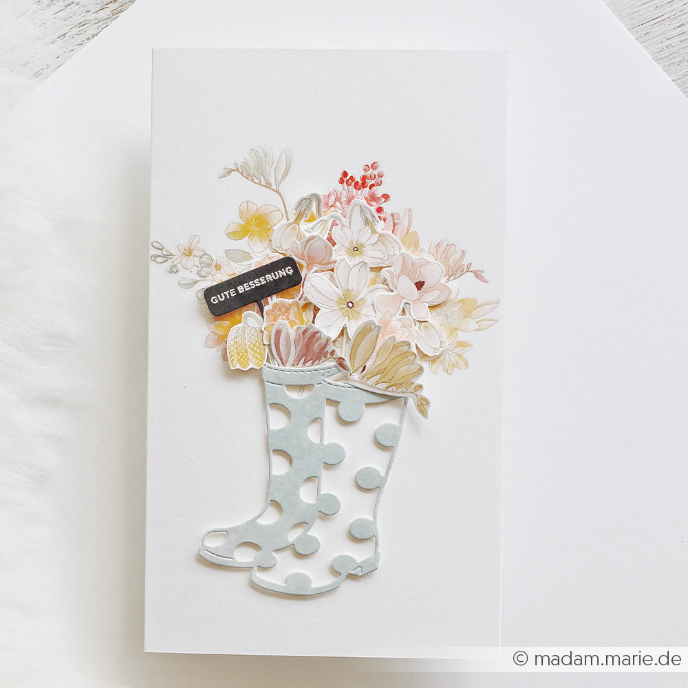 Karte mit gepunkteten Gummistiefeln, gestaltet mit der Motivstanze „Gummistiefel“ von Alexandra Renke, verziert mit pastellfarbenen Blumen und einem Anhänger mit der Aufschrift „Gute Besserung“. Ideal zum Basteln mit Stanzen für Designpapier in Ihren Projekten.