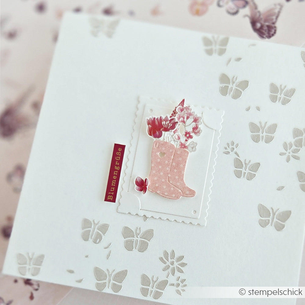 Weiße Karte mit rosa Stiefeln und Blumen, Schmetterlingsausschnitten aus dem „Gartenfreuden“-Set von Alexandra Renke sowie dem Schriftzug „Blumengrüße“ auf der Vorderseite.