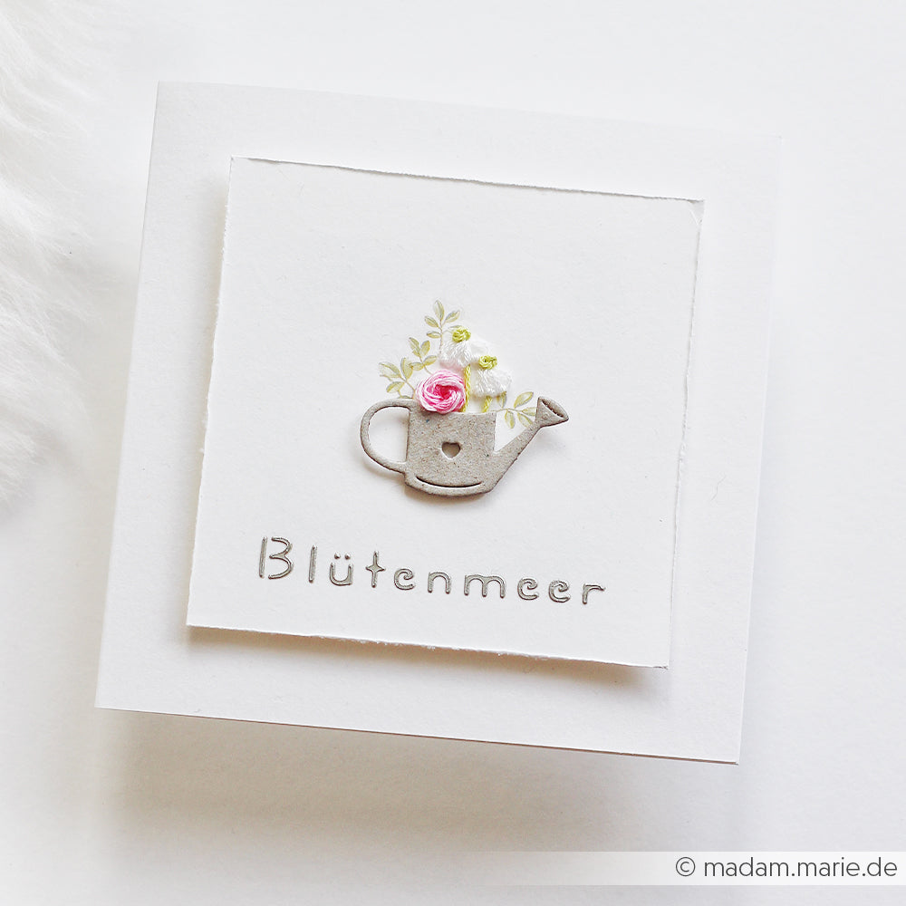 Eine weiße Karte von Alexandra Renke mit einer Illustration einer Gießkanne und Blumen, die in Grau mit „Blütenmeer“ beschriftet ist und mit Designpapieren sowie der Motivstanze „Gartenfreuden“ für detailreiche Akzente gestaltet wurde.