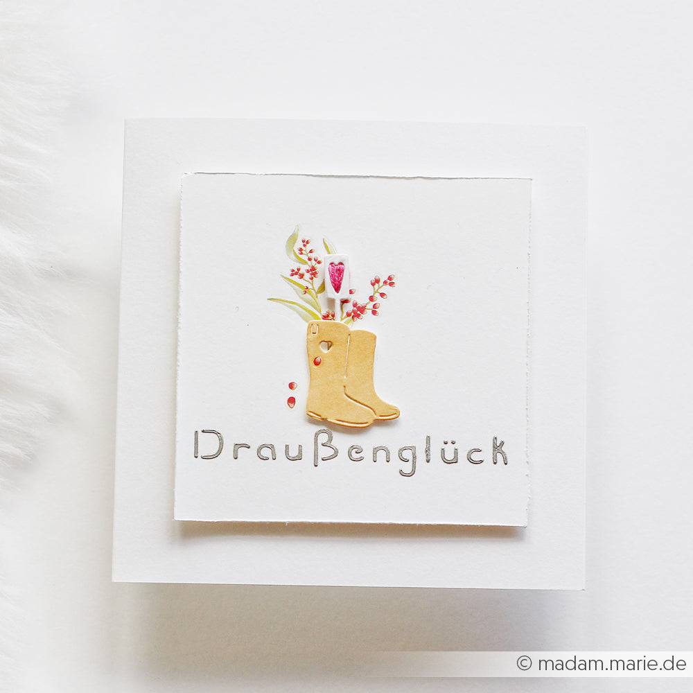 Quadratische Karte von Alexandra Renke mit einer Illustration von gelben Stiefeln, die Blumen über dem grauen Schriftzug „Draußenglück“ halten – ideal für Fans der Motivstanze „Gartenfreuden“ und kreativer Projekte mit Designpapieren.