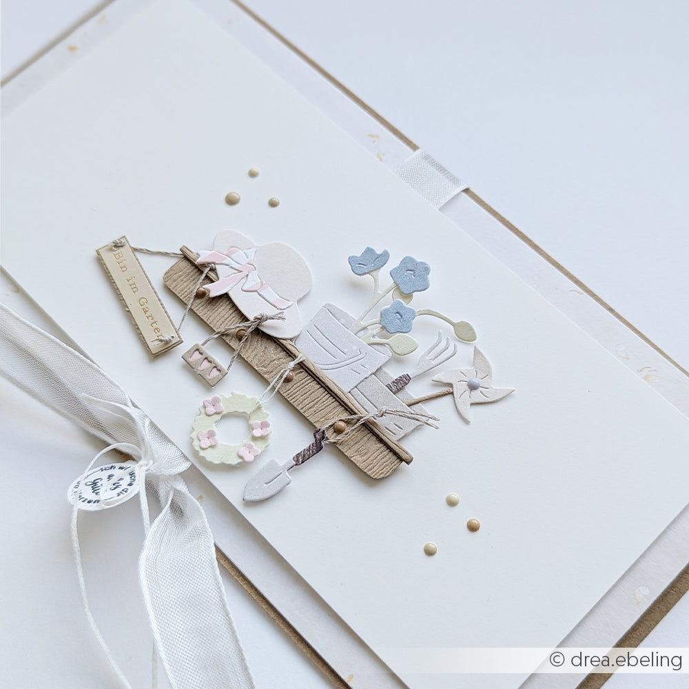 Handgefertigte Karte mit mehrschichtigen Papierausschnitten, gestanzt mit den „Stanzen für Designpapiere“ von Alexandra Renke, mit einem Zaun, Blumen, Werkzeugen und einem Schild mit der Aufschrift „Pflanzen machen gute Laune“ – perfekt für jede Kollektion der „Motivstanze Gartenspaß Anhänger“.
