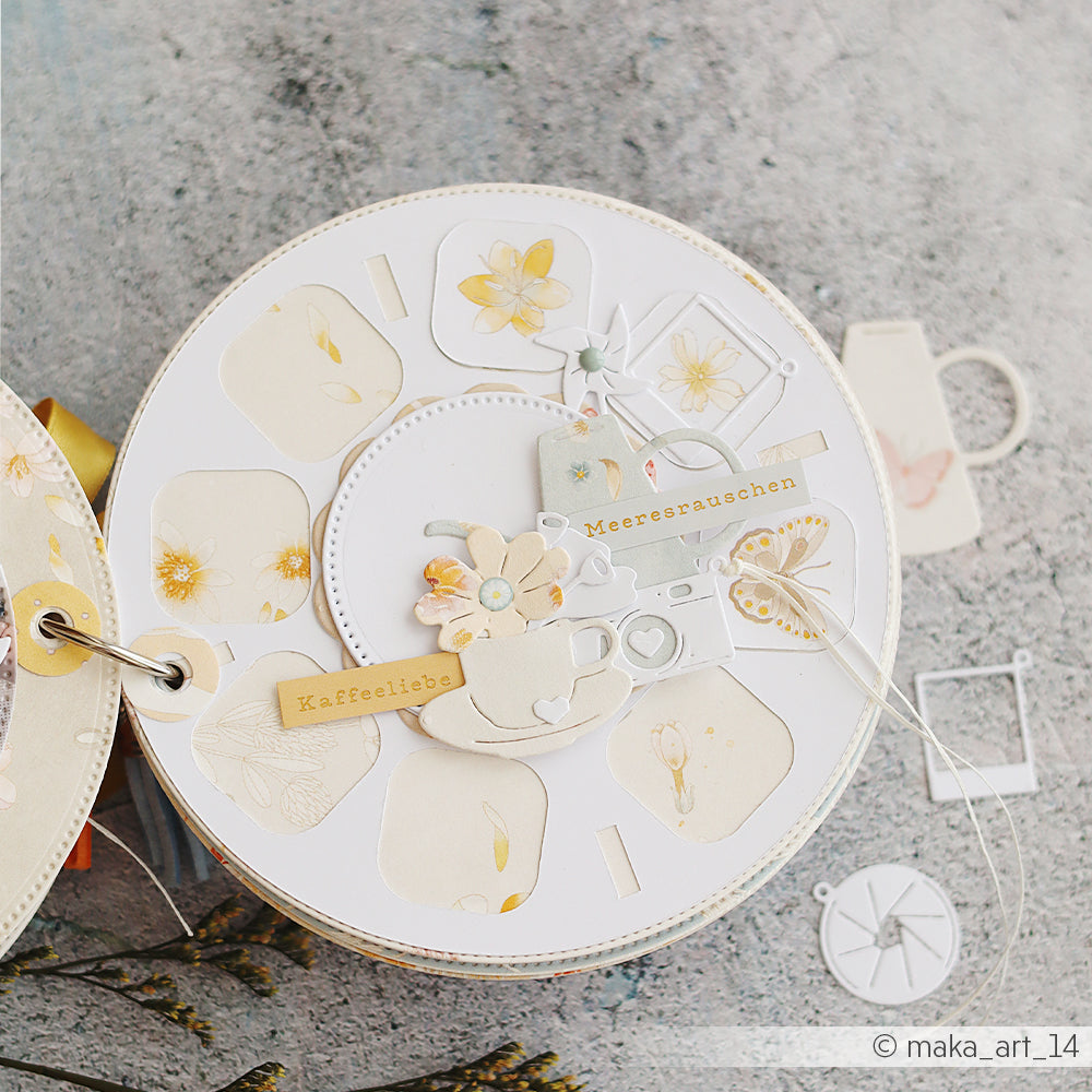 Eine runde Scrapbook-Seite mit pastellfarbenen Blumenmotiven, ausgestanzten Teetassen und Wasserkochern, die mit dem DieCut-System von Alexandra Renke hergestellt wurden, sowie Verzierungen mit deutschen Wörtern auf hochwertigen Designpapieren.