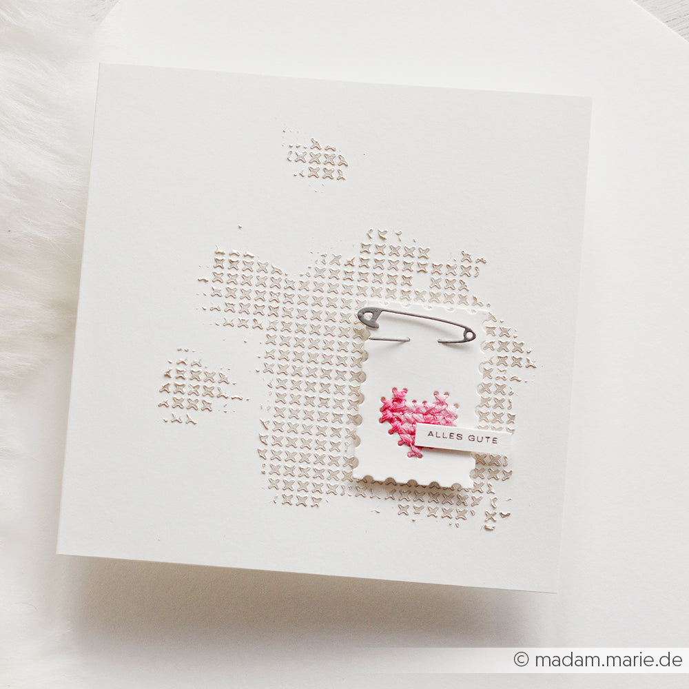 Weiße Karte von Alexandra Renke mit rosa Kreuzstichherz, Sicherheitsnadel und kleinem „ALLES GUTE“-Etikett, gestaltet mit „Stanzen für Designpapier“ auf strukturiertem Hintergrund.