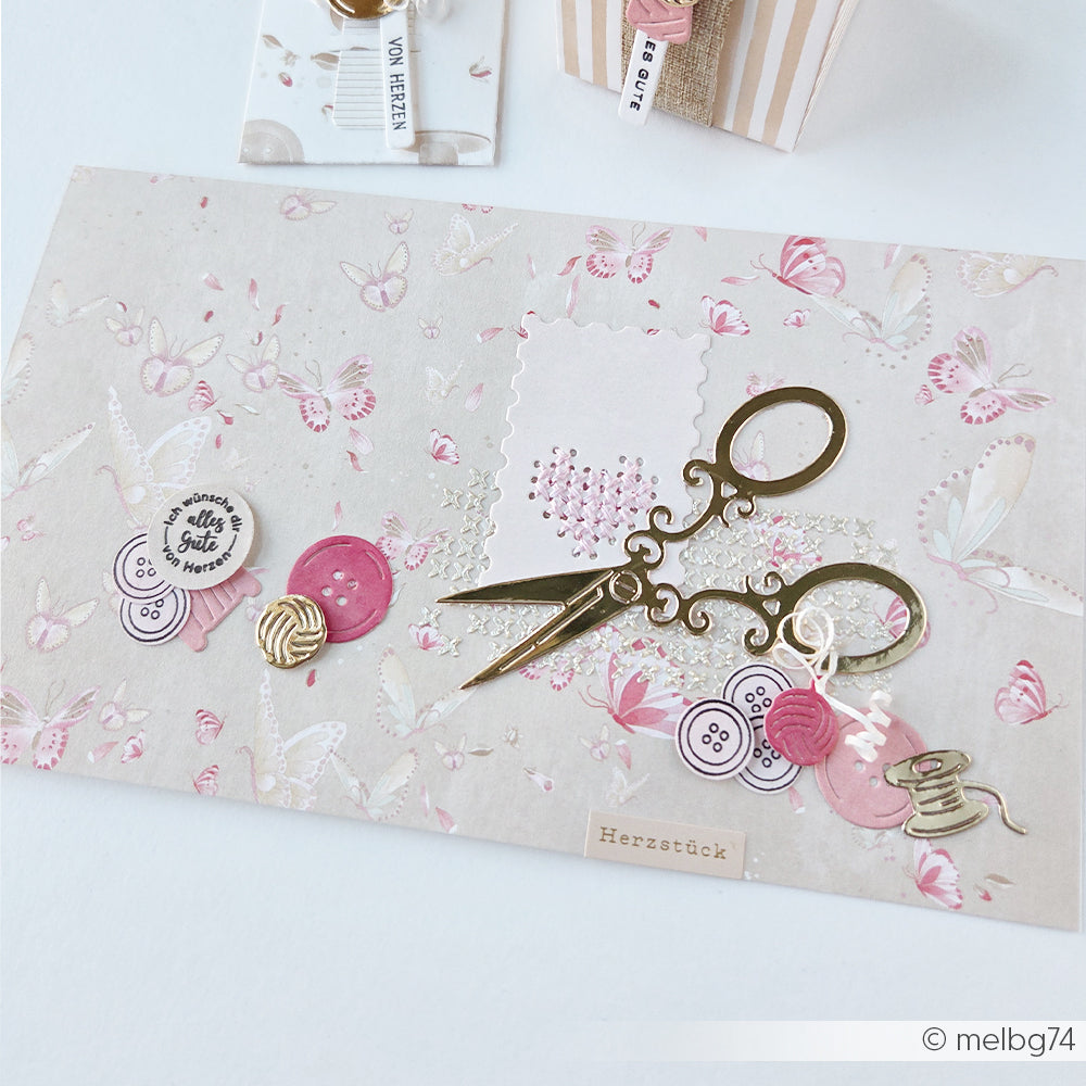 Eine dekorative Karte mit goldenen Scheren, Knöpfen und rosa Akzenten vor einem Schmetterlingshintergrund, gefertigt mit den „Stanzen für Papier“ von Alexandra Renke für präzise Formen und Details.