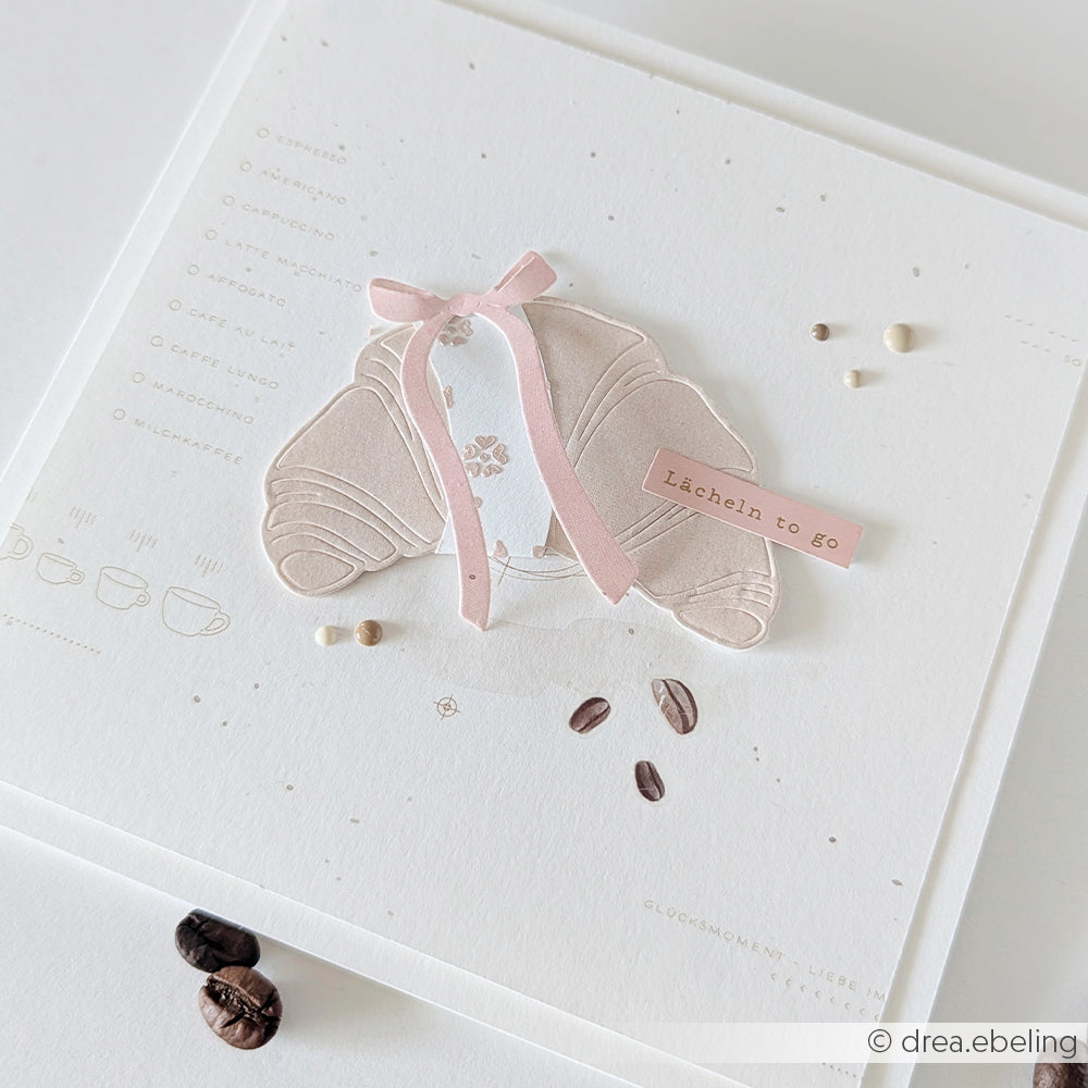 Eine minimalistische Karte von Alexandra Renke zeigt eine Croissant-Illustration, die mit der Motivstanze „Croissant Outline“ gestaltet und mit einem rosa Band, Kaffeebohnen und dem Schriftzug „Lächeln to go“ verziert wurde – perfekt zum Kombinieren mit Ihren Lieblings-Designpapieren.