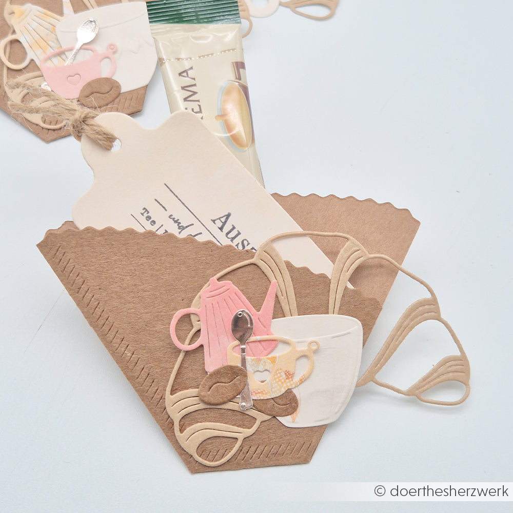 Brauner Papiertütchen, verziert mit ausgestanzten Motiven wie Kaffeetassen, einer Kaffeekanne, Kaffeebohnen, einem Anhänger und einem kleinen Kaffeesäckchen – gefertigt aus hochwertigen Designpapieren und mit Stanzsystemen von Alexandra Renke für präzise Details. Zu den Besonderheiten zählen charmante Akzente wie die Motivstanze „Croissant Outline“ von Alexandra Renke, die dem Ganzen eine besonders verspielte Note verleihen.