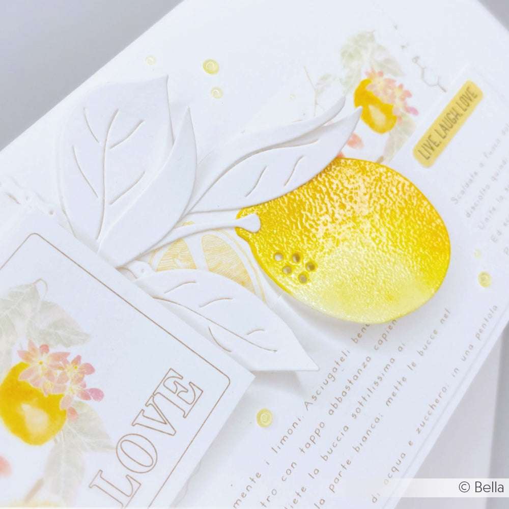 Weiße Karte mit geprägten Blättern, einer gelben Zitrone aus der Zitrone-Stanzform von Alexandra Renke und dem Wort LOVE auf einem Anhänger, der mit hochwertigem Designpapier von Alexandra Renke hergestellt wurde und einen eleganten Abschluss bildet.