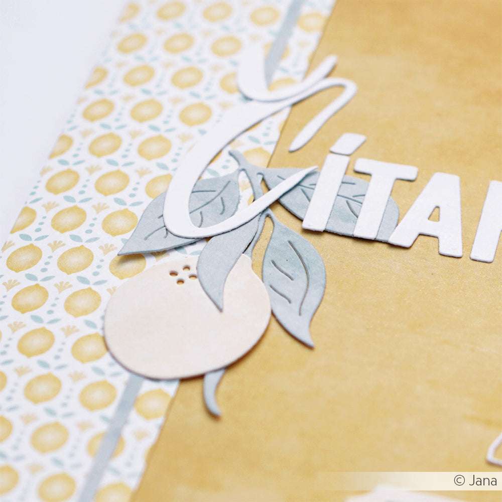 Nahaufnahme einer dekorativen Papierarbeit mit ausgeschnittenen Buchstaben, Blättern und Zitronen, die mit der Alexandra Renke Motivstanze Zitrone auf einem gelb-weiß gemusterten Hintergrund hergestellt wurde.