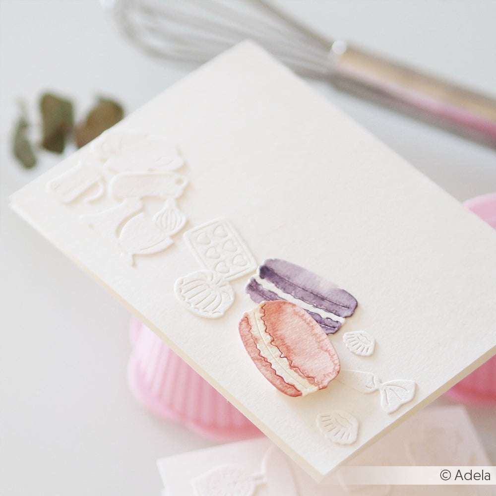 Eine handgefertigte Karte von Alexandra Renke mit geprägten Küchenwerkzeugen und aquarellierten Macarons auf weißem Designpapier, verziert mit einer Motivstanze Macarons für zusätzlichen Charme.