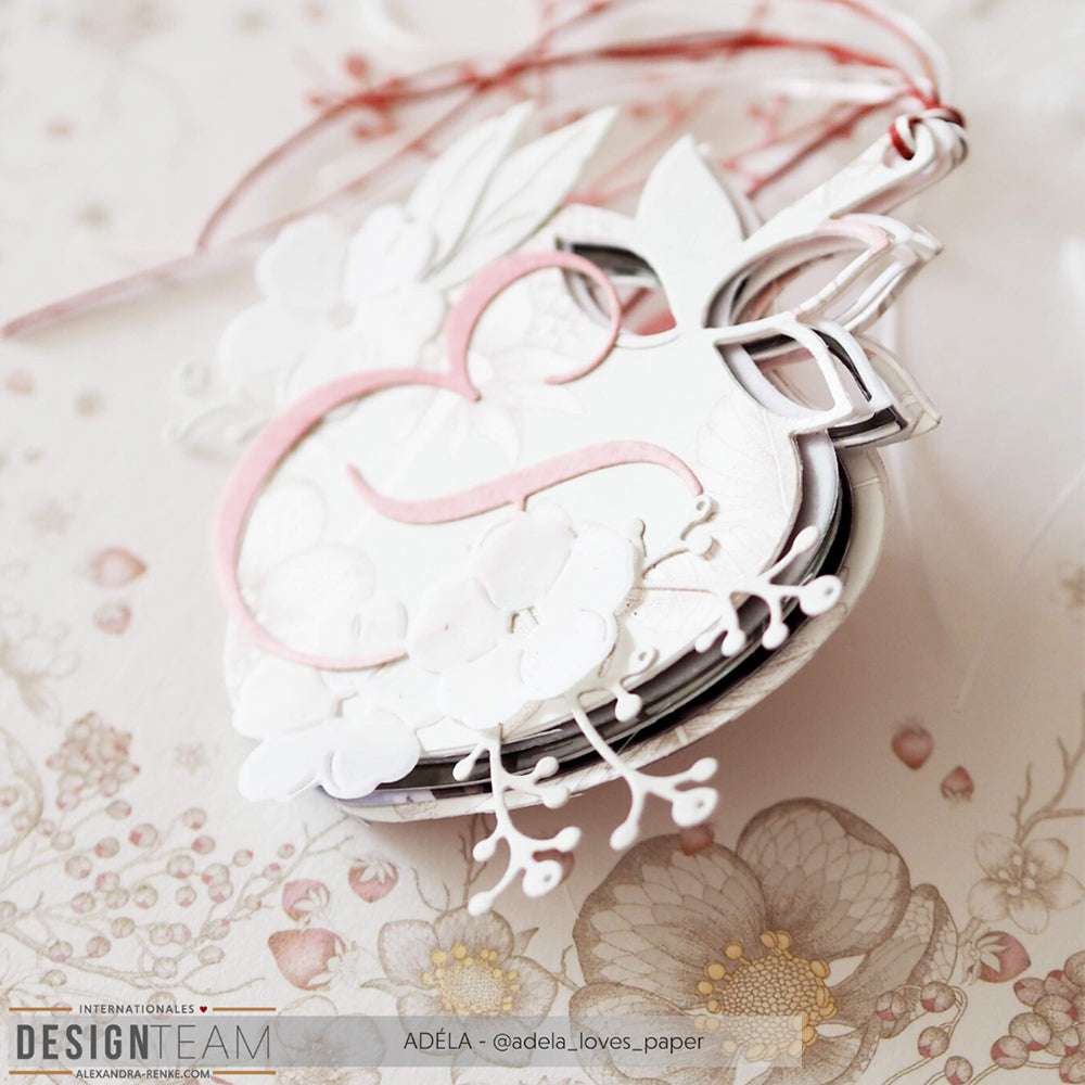 Nahaufnahme eines zarten, geschichteten Blumenornaments aus Alexandra Renke Designpapieren in Rosa und Weiß vor einem floralen Hintergrund, das die filigranen Ergebnisse verdeutlicht, die mit DieCut Systemen möglich sind.
