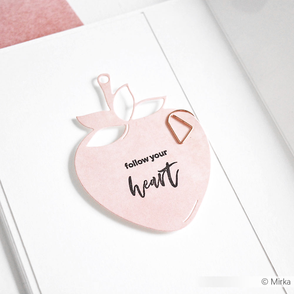 Eine rosafarbene, erdbeerförmige Karte, die mit der Alexandra Renke Motivstanze Erdbeer Etikett mit dem Text "follow your heart" gestaltet wurde, wird mit einer Büroklammer auf eine weiße Fläche gelegt.