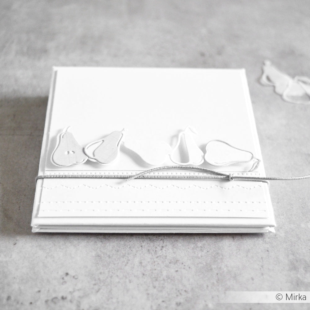 Weiße Karte mit geprägten Birnen aus der Alexandra Renke Motivstanze Birnenreihe, verziert mit einer silbernen Schnur, auf einer grauen Fläche.