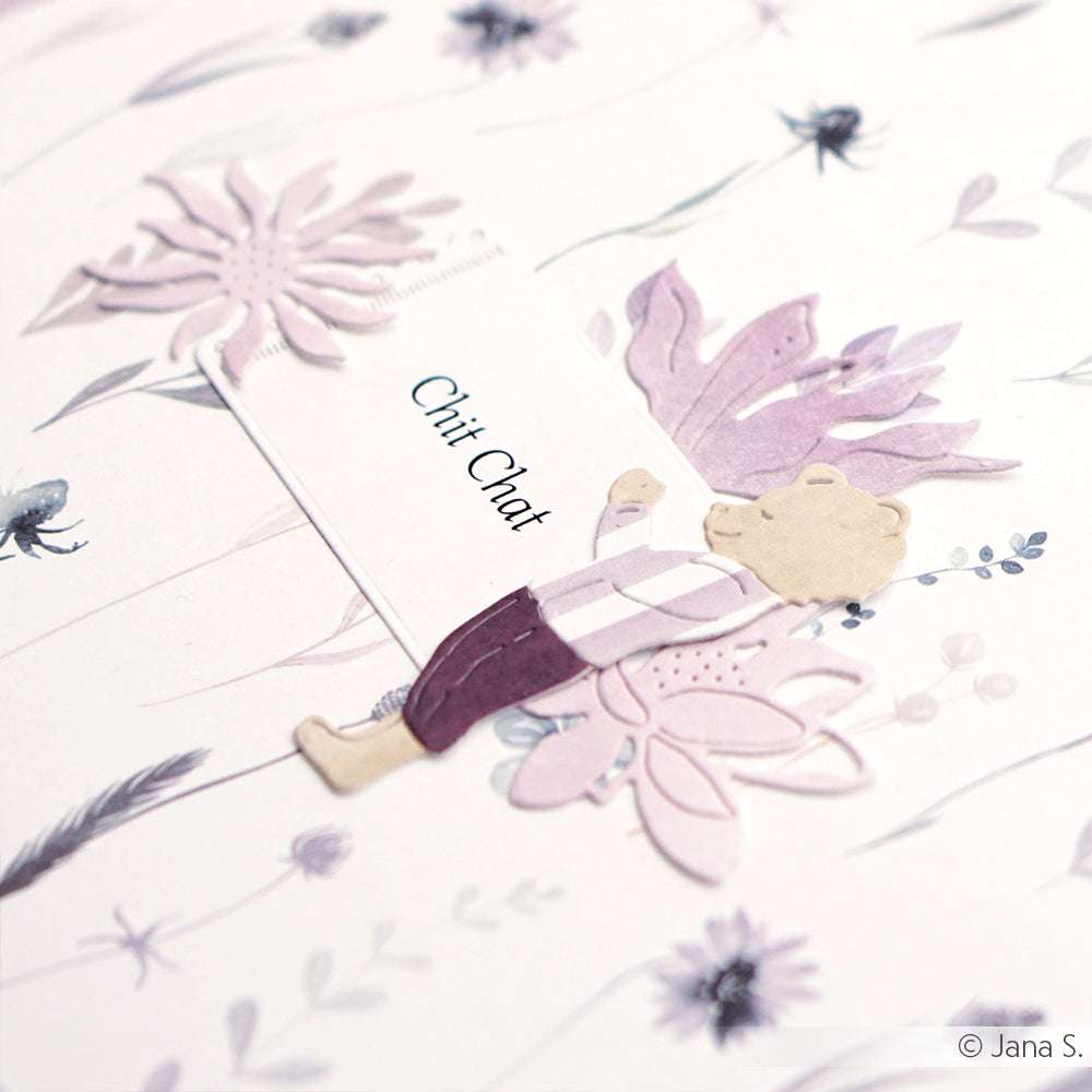 Ein Bär in lila Kleidung hält eine "Chit Chat"-Karte, die mit den Motivstanze Blumenpaar von Alexandra Renke vor einem floral gemusterten Hintergrund gestaltet wurde.