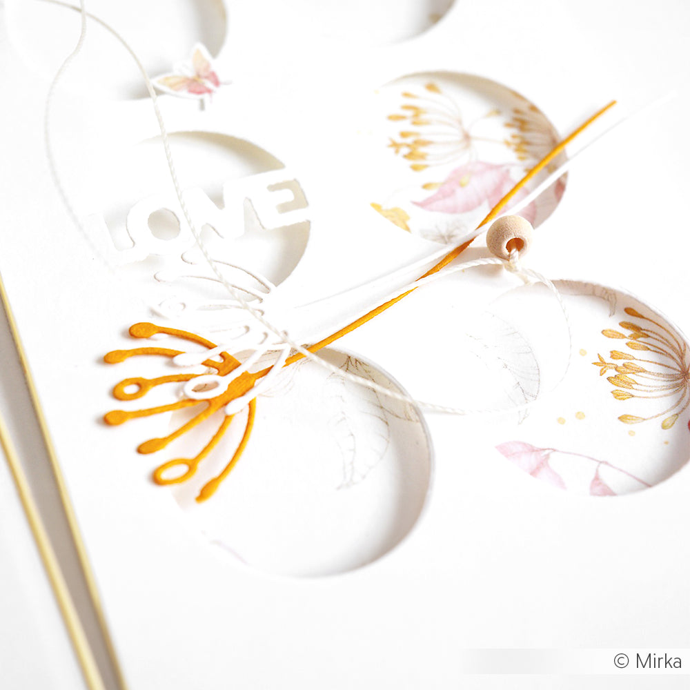Weiße Karte mit kreisförmigen Ausschnitten und floralen Motiven, die mit dem Artistblumen-Set und der Motivstanze von Alexandra Renke hergestellt wurden, mit dem Text LOVE und einer Schnur mit einer kleinen Holzperle.