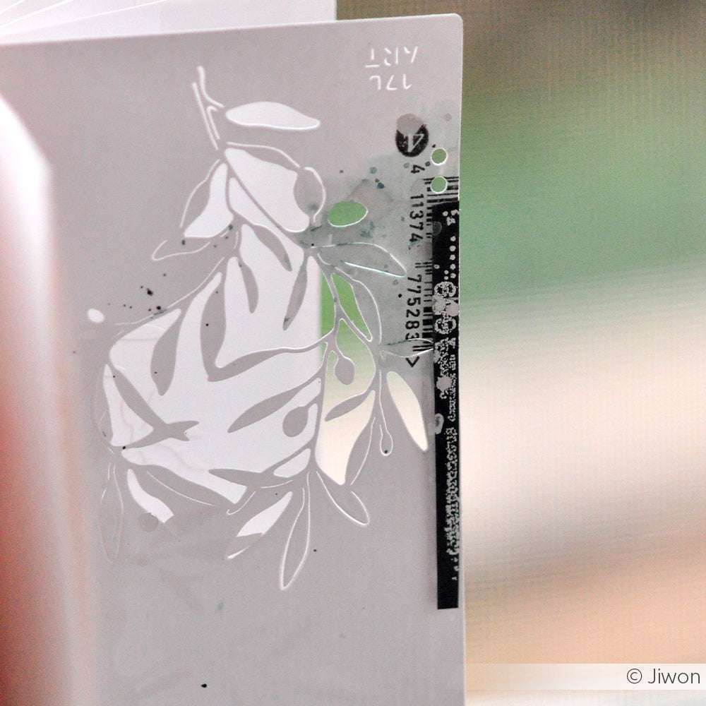 Eine weiße Karte mit einem Blattausschnitt aus der Alexandra Renke Motivstanze Olivenzweig und einem schwarz gedruckten Text, der sich vor einem verschwommenen Hintergrund abhebt.