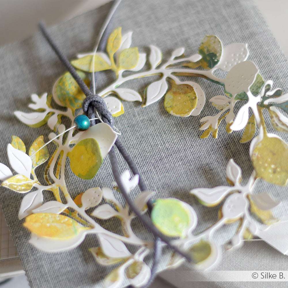 Das Geschenk ist in grauen Stoff eingewickelt, mit einem Blumenkranz aus Papier verziert, der mit Alexandra Renke Motivstanze Zitronenkranz Dies hergestellt wurde, und mit einer Schnur mit einer tealfarbenen Perle versehen.