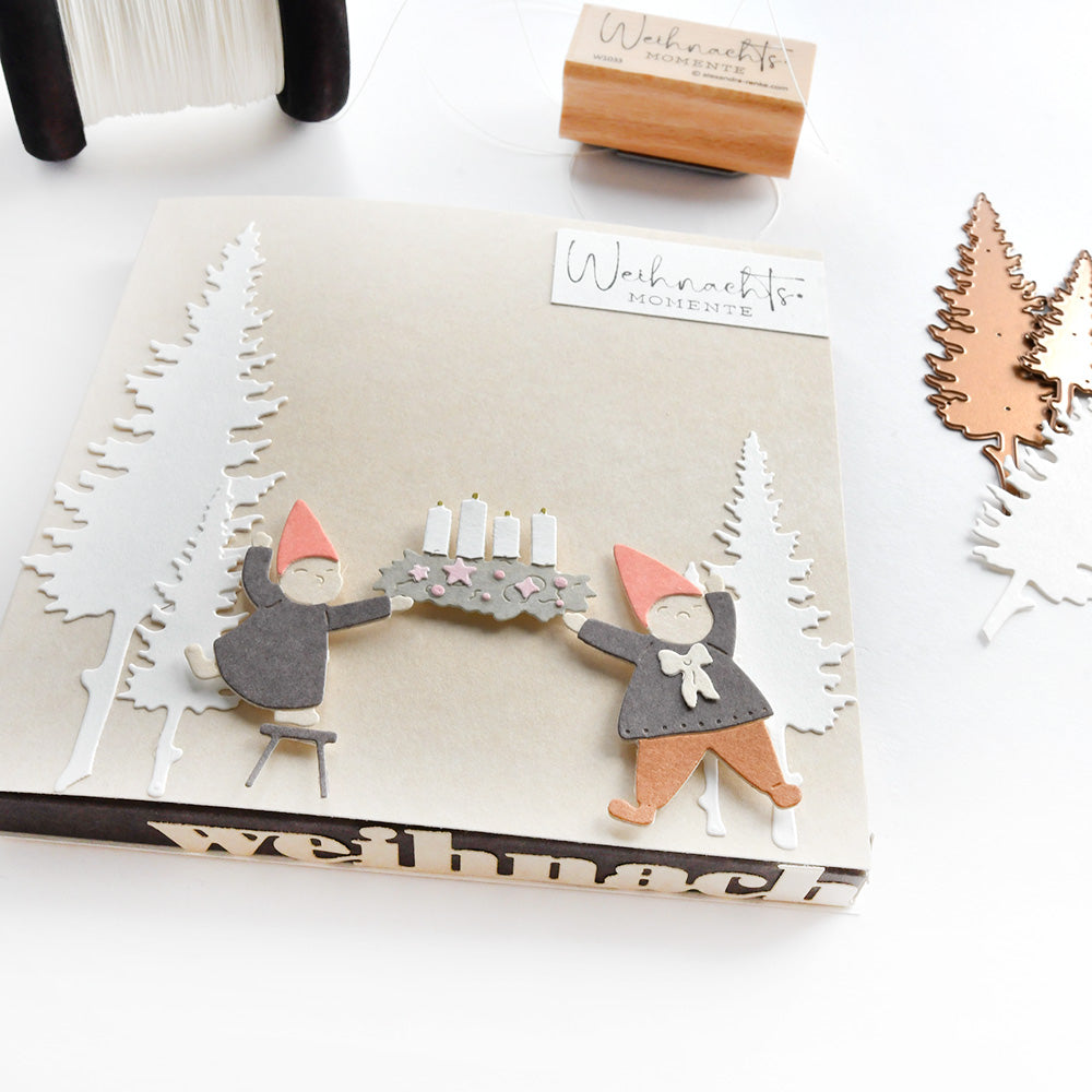 Handgefertigte Weihnachtskarte von Alexandra Renke mit zwei Zwergen, die einen Kerzenkranz halten, festlichen Ausschnitten, Bäumen aus Designpapieren und detaillierten Akzenten aus der Motivstanze Fünf Tannen Stanzsysteme.