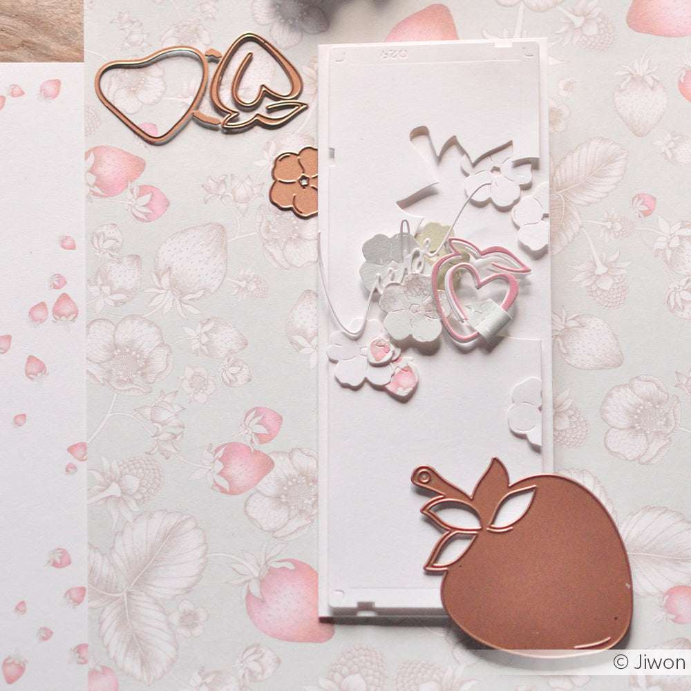 Erdbeerförmige Papier- und Metallausschnitte, die mit Alexandra Renke Stanzen hergestellt wurden, sind auf blumengemustertem Papier in zarten Rosatönen angeordnet.