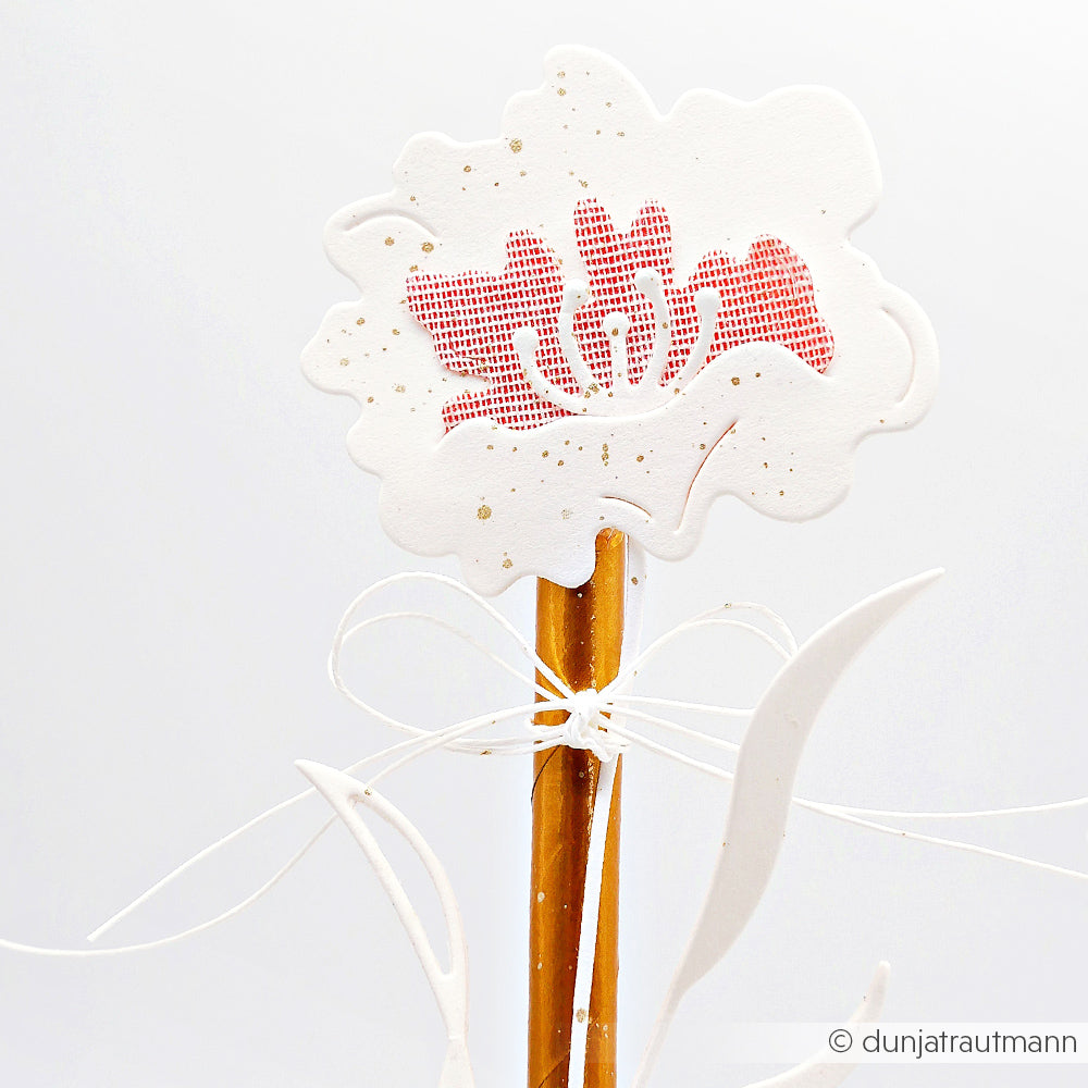 Weiße Papierblume mit rot gemusterter Mitte, goldenem Stiel und weißer Schleife auf einfarbigem Hintergrund - hergestellt mit Alexandra Renke DieCut Systemen und Motivstanze Schichtblume für komplizierte Designdetails.