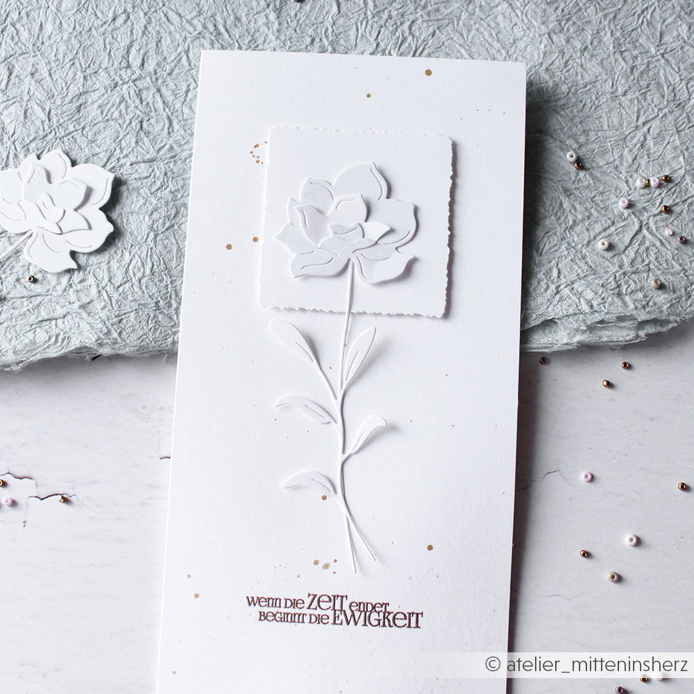 Weiße Karte mit geprägtem Blumenmuster aus der Alexandra Renke Schichtblume 3 Motivstanze und deutschem Text, auf grauem Strukturpapier mit verstreuten Perlen.