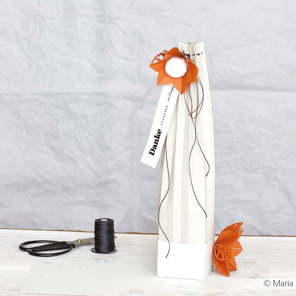 Große Geschenktüte mit orangefarbener Papierblume aus Alexandra Renke Motivstanze Faltblume, schwarzer Schnur und "Danke"-Anhänger; daneben eine Spule mit schwarzem Garn.