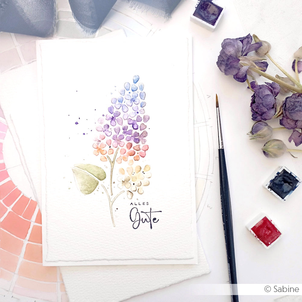 Aquarell einer lila Blume auf 200g weißem Designpapier von Alexandra Renke, mit Pinsel, getrockneten Blumen und Farben in der Nähe; ideal für die Verwendung mit Motivstanze Negativ-Blume 2 oder DieCut Systemen.