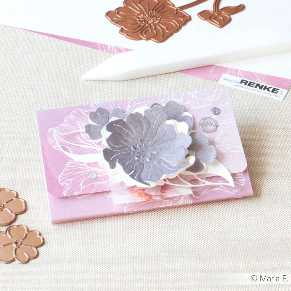 Ein rosafarbener Umschlag von Alexandra Renke, verziert mit geschichteten Papierblumen, floralen Stanzungen und Akzenten der Motivstanze Negativ-Blume 4, ausgestellt auf einer leichten Stoffoberfläche.