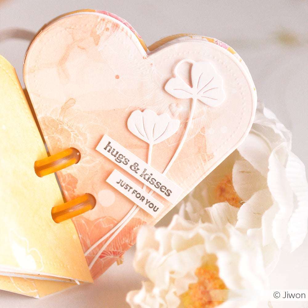 Pastellfarbene Karte in Herzform mit floralen Motiven, die mit den Stanzformen von Alexandra Renke hergestellt wurde und den Text "hugs & kisses just for you" neben weißen Blumen zeigt.