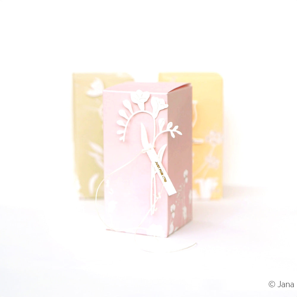 Eine rosafarbene Geschenkbox mit floralem Ausschnitt, gefertigt aus der Alexandra Renke Motivstanze Glockenblümchen, steht vor zwei pastellfarbenen Notizbüchern aus den hochwertigen Designpapieren von Alexandra Renke.