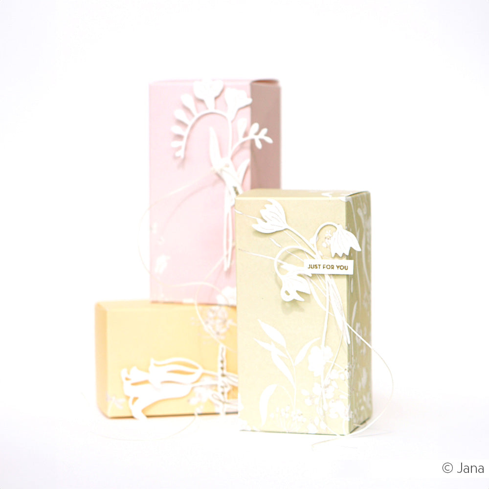 Drei pastellfarbene Geschenkschachteln von Alexandra Renke mit zarten weißen Blumenmustern aus Papier, die mit Stanzformen hergestellt wurden, jeweils mit einem Anhänger auf weißem Hintergrund.