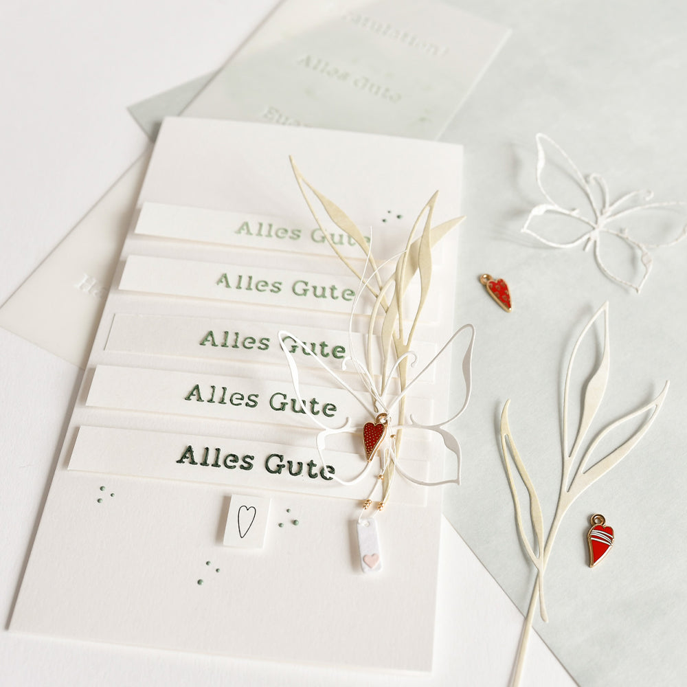 Mehrere Alles Gute Karten mit Papierausschnitten, getrocknetem Gras und Herzanhängern auf einer weißen Oberfläche, hergestellt mit Alexandra Renke Motivstanze Lineart Blattwerk und eleganten Designpapieren für eine einzigartige handgemachte Note.