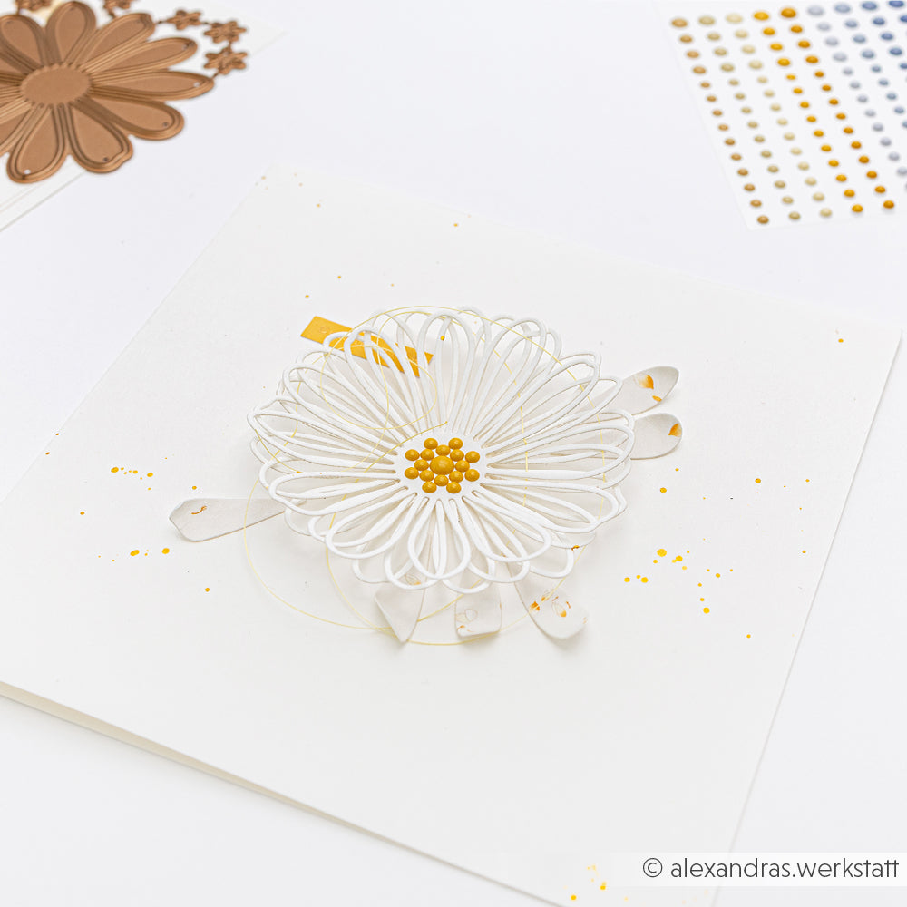 Eine handgefertigte Karte mit einer weißen Papierblume mit gelber Mitte, hergestellt mit Alexandra Renke Designpapieren und der Motivstanze Margerite Outline, umgeben von buntem Bastelmaterial.