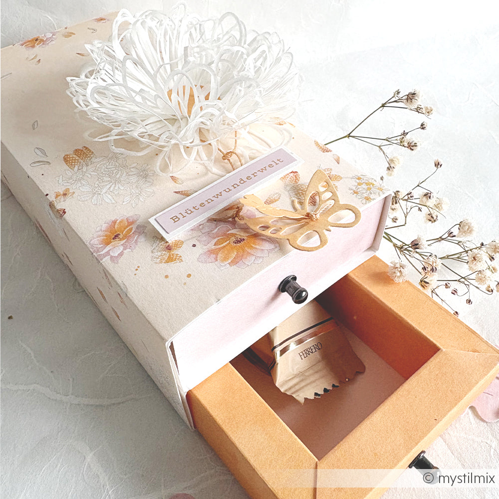 Geschenkbox mit Blumenmotiv von Alexandra Renke, verziert mit einer weißen Papierblume, die mit der Motivstanze „Margerite Outline“ gefertigt wurde, einem Schmetterlingsausschnitt und einer Schublade, in der ein kleines Holzobjekt Platz findet – ideal zur Präsentation Ihrer Lieblingsprodukte von DieCut Systeme.