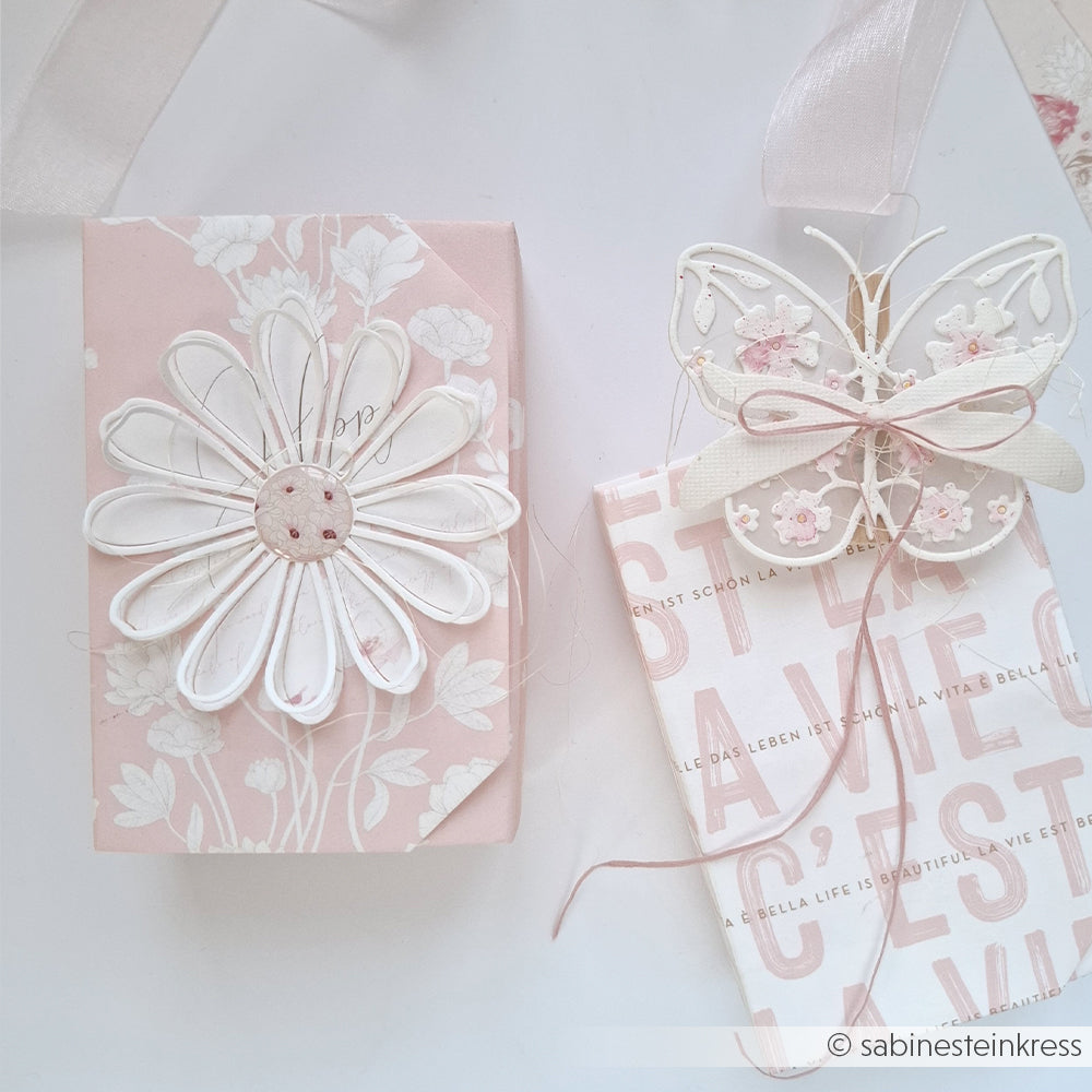 Ein Geschenk mit einer Margerite und einer Karte mit Schmetterling, beide in zartem Rosa und Weiß, gestaltet mit der Motivstanze „Margerite Outline“ von Alexandra Renke und eleganten Designpapieren, vor einem hellen Hintergrund.
