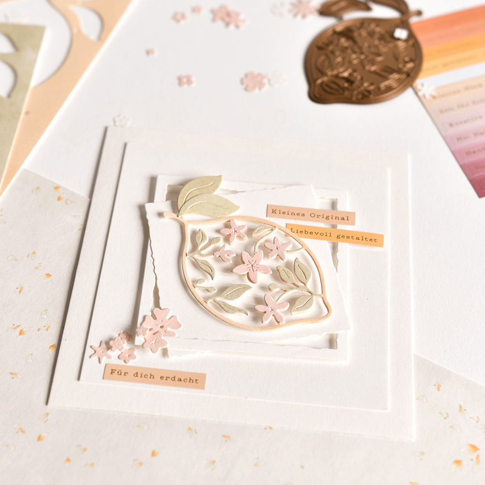 Eine handgefertigte Karte von Alexandra Renke mit zarten rosa und beigen Blumenmustern auf hochwertigem Designpapier, hergestellt mit der Motivstanze Blumige Zitone und deutschen Etiketten, vor weißem, strukturiertem Hintergrund.