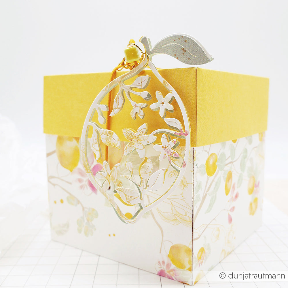 Dekorative Geschenkbox zum Thema Zitrone von Alexandra Renke mit einem gelben Deckel und einem bezaubernden „Blumige Zitone“-Anhänger, der mit hochwertigen Stanzsystemen gefertigt und oben angebracht ist.