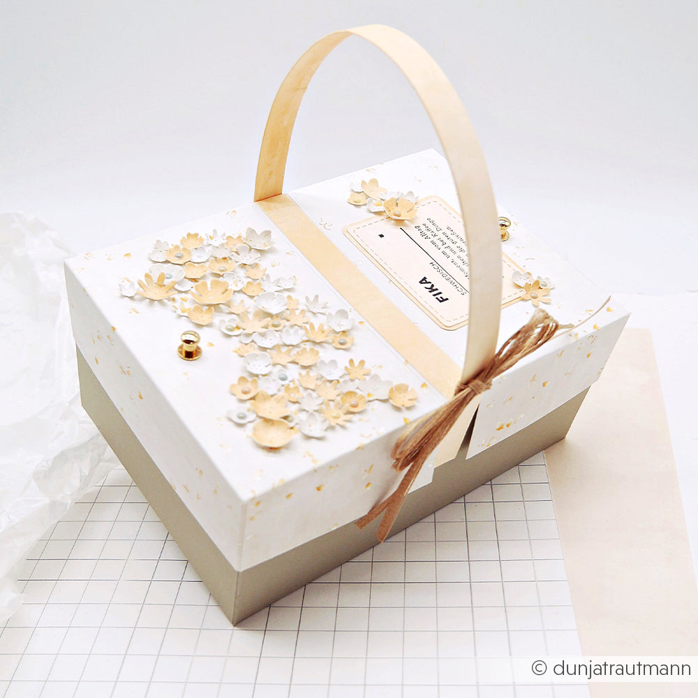 Dekorative Geschenkbox mit Griff und floralen Akzenten, gestaltet mit der Motivstanze „Streublümchen“ von Alexandra Renke und verziert mit einer Schnurschleife auf Karopapier. Ideal, um Ihre Lieblings-Designpapiere stilvoll zu verpacken.