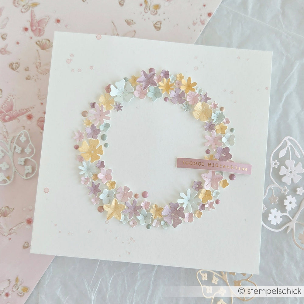 Eine quadratische Karte von Alexandra Renke mit einem pastellfarbenen Blumenkranz, der mit der Motivstanze „Streublümchen“ gestaltet wurde, und einem kleinen rosa Anhänger mit der Aufschrift „10000 Bittergrüße“.