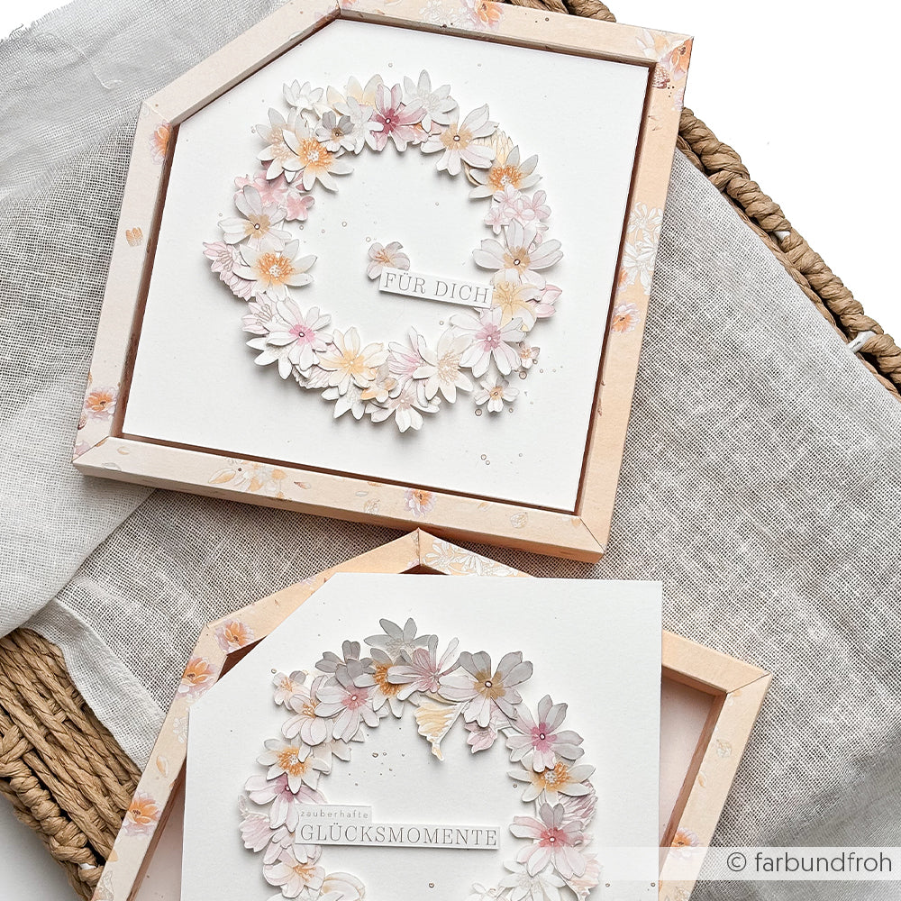 Zwei quadratische Karten von Alexandra Renke mit pastellfarbenen Blumenkränzen und deutschem Text, gefertigt aus hochwertigen Designpapieren und dem Stanzsystem „Streublümchen“, sind in Rahmen mit rosa Rand auf Stoff präsentiert.